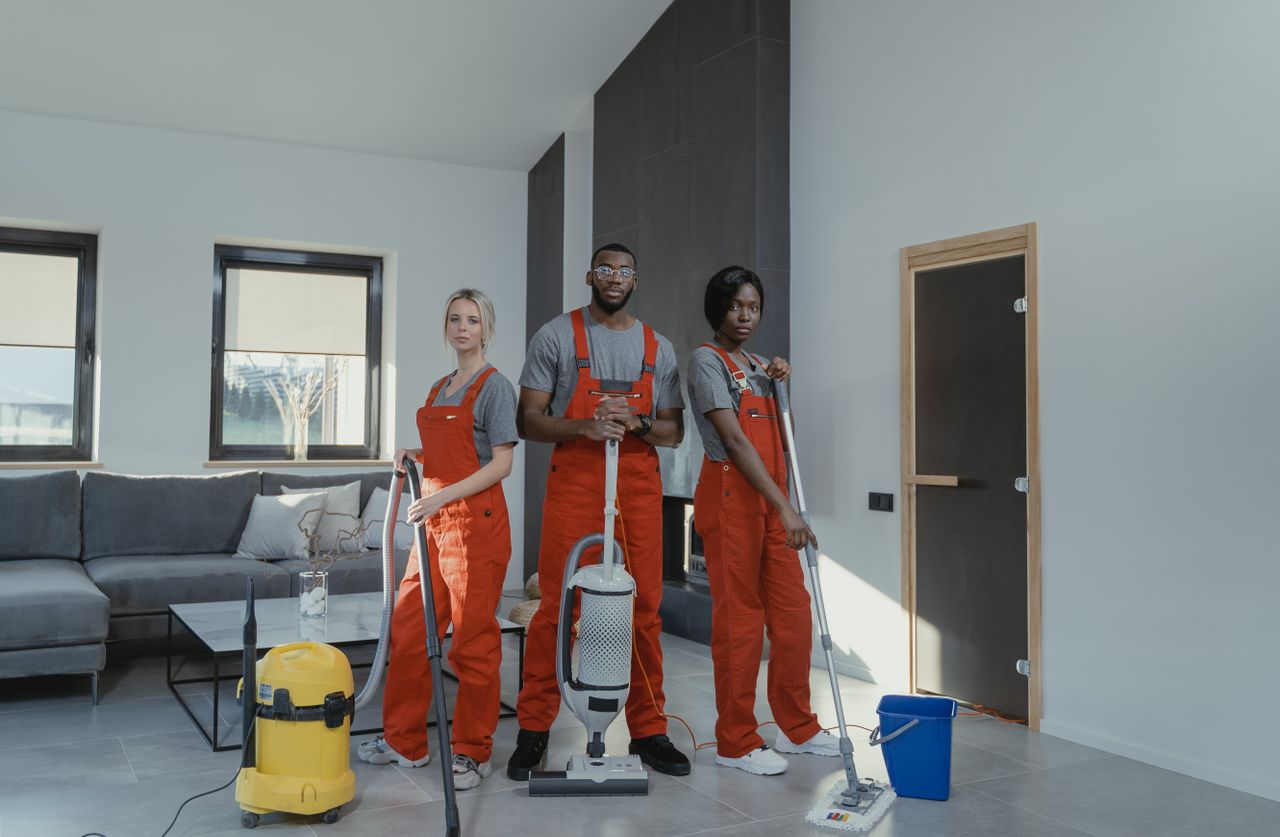 services-02 Diverse cleaning team in red uniforms ready to sanitize a modern living room with vacuum and mop.
